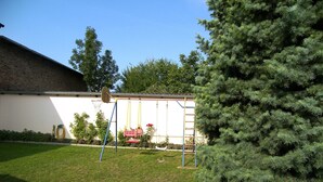 Ein Garten mit Schaukel, Basketballkorb, einer hohen Nadelbaum und einem weißen Gebäude mit Backsteinmauer.
