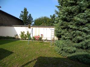 A backyard with a swing set, a basketball hoop, a tall evergreen tree, and a white building with a brick wall.