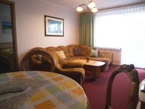 A living room with a sofa, a coffee table, and a round table with a checkered tablecloth. There is a framed picture on the wall and a window with curtains.