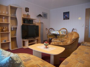 Ein Wohnzimmer mit Fernseher, Couchtisch, Sofa und Esstisch mit Stühlen.
