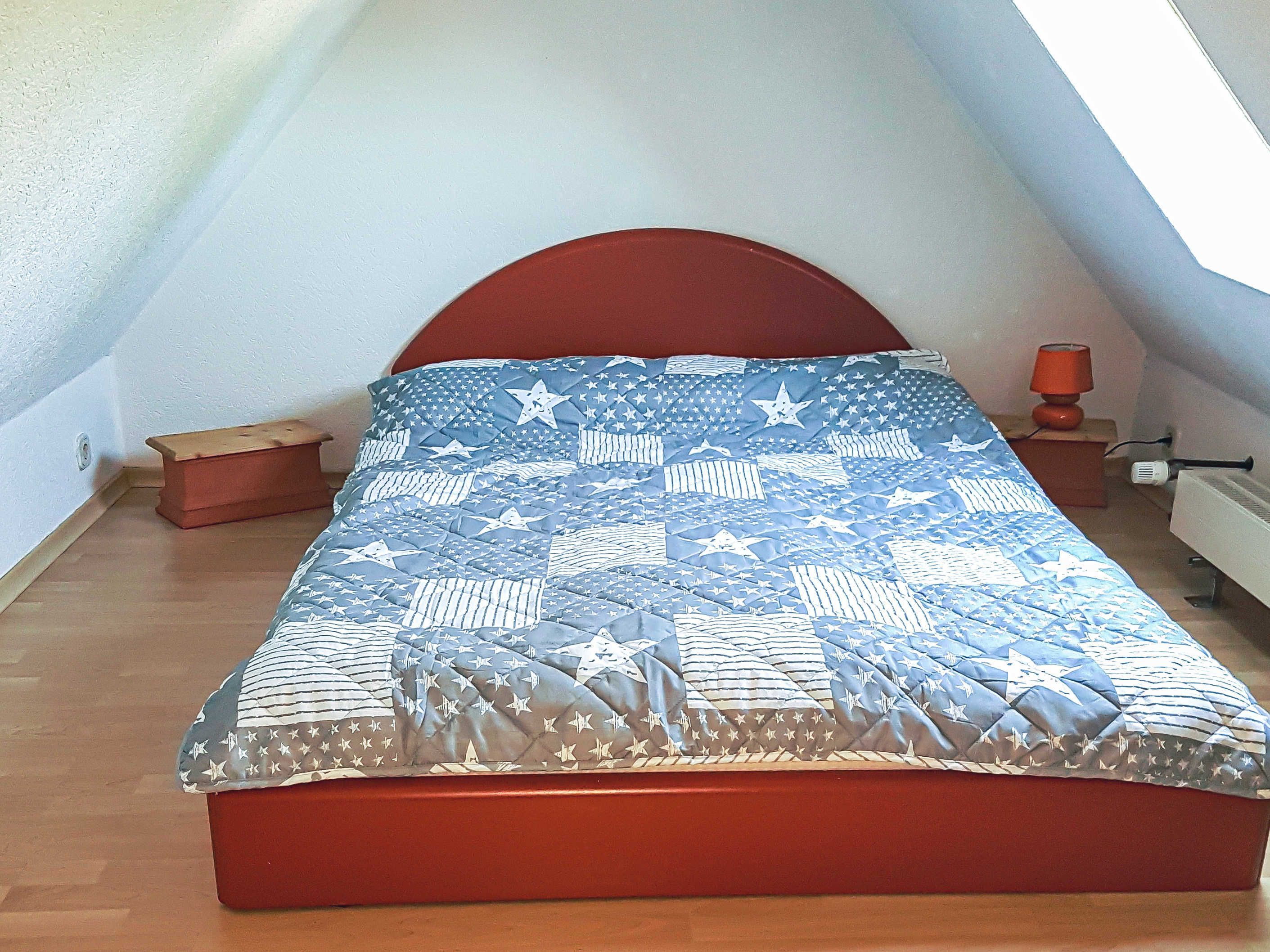 Un dormitorio con techo inclinado, un cabecero rojo, una colcha con diseño de estrellas, un taburete de madera pequeño y una mesita de noche con lámpara.