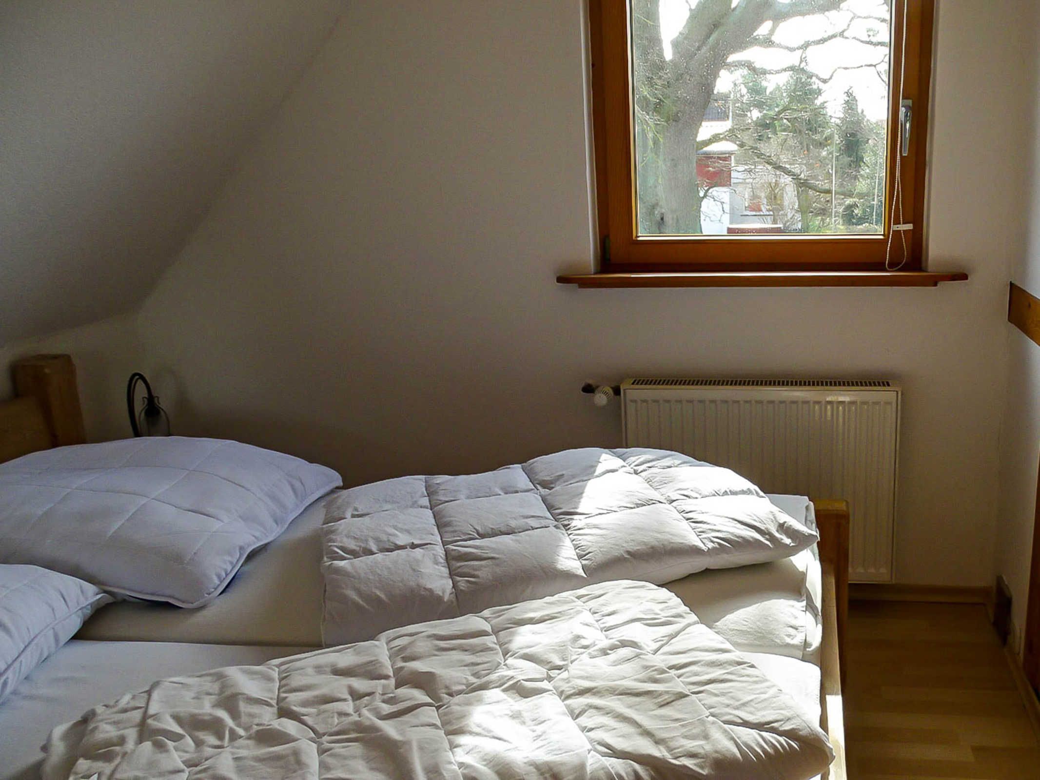 Un dormitorio con techo inclinado, una ventana con vistas a árboles y edificios, un radiador y una cama con ropa de cama blanca.