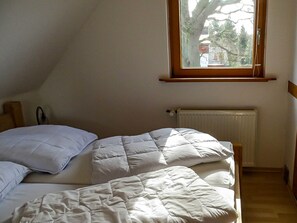 A bedroom with a sloped ceiling, a window with a view of trees and buildings, a radiator, and a bed with white bedding.