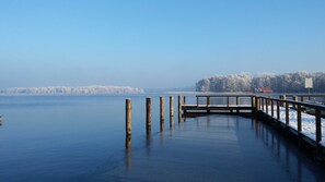 Un lac gelé avec une jetée en bois qui s’avance sur l’eau, entouré d’arbres couverts de neige et sous un ciel dégagé.