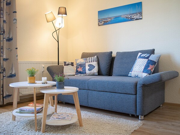 Un salon chaleureux avec un canapé gris, une petite table ronde surmontée d’une lampe et un tableau représentant un port de plaisance accroché au mur.