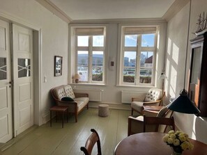 Une pièce offrant une vue sur le paysage urbain grâce à de grandes fenêtres, une table ronde agrémentée d’un vase de fleurs et un coin salon confortable avec un canapé et des fauteuils.