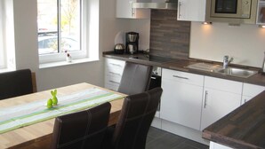 A modern kitchen with white cabinets, a stainless steel microwave, a built-in oven, and a window with blinds. - 3-room 104 Jordland Balk Spülm. (BC 5) - BUE - Apartment house Westwind (Büsum)