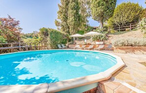Una piscina con sillas de descanso y sombrillas, rodeada de árboles y un patio de piedra.