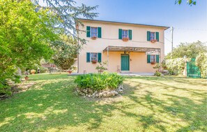 Una casa de dos pisos con una puerta verde, varias ventanas con contraventanas verdes y un pequeño jardín con un muro de piedra y plantas en macetas.