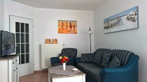 Een woonkamer met een televisie, een bank, een salontafel met een bloevas en twee schilderijen aan de muur.