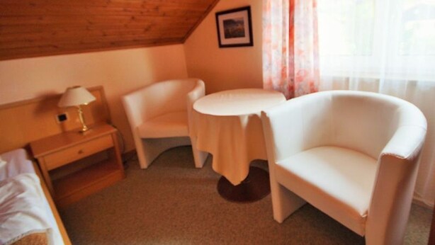 Une petite pièce chaleureuse avec un plafond en bois, un lit, une table de chevet avec une lampe, un fauteuil blanc, une table ronde et une fenêtre avec des rideaux.