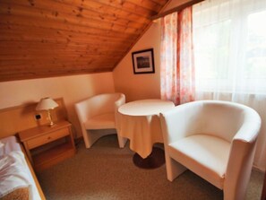 Ein kleiner, gemütlicher Raum mit Holzboden, einem Bett, einem Nachttisch mit Lampe, einem weißen Sessel, einem runden Tisch und einem Fenster mit Vorhängen.