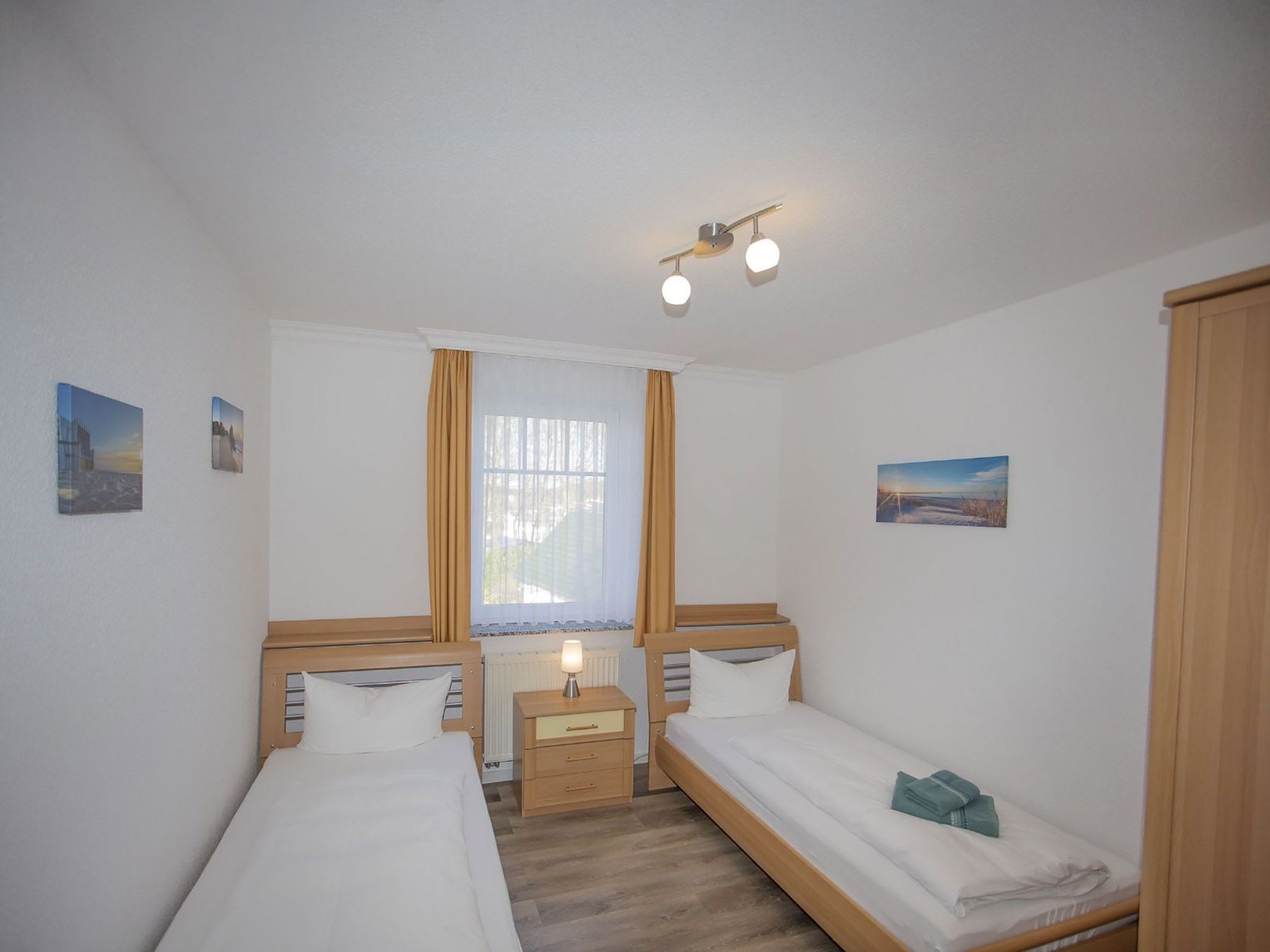 A room with two single beds, a window with curtains, a nightstand with a lamp, and wall art.