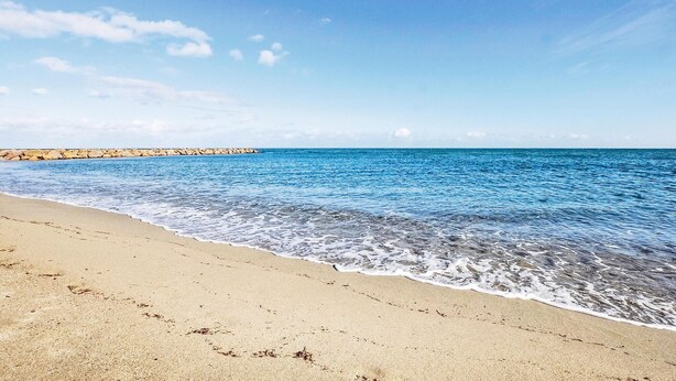 Una playa con agua azul cristalina, una orilla de arena y un rompeolas rocoso a lo lejos.