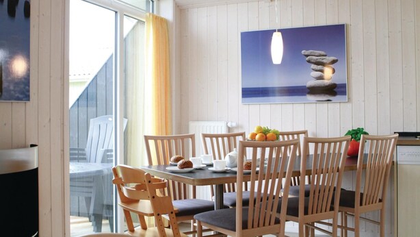 A dining area with wooden chairs and a table set for a meal, featuring a painting of a rock formation and a view of the sea. - Friedrichskoog-Deichblick 7 (Friedrichskoog)