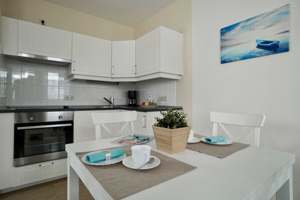 A modern kitchen with white cabinetry, a built-in oven, and a coffee maker. There is a dining table with a plant and a magazine.