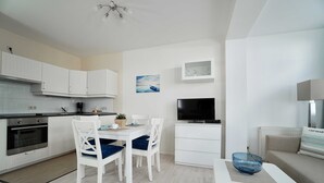A modern kitchen with white cabinets, a dining table with blue chairs, a TV, and a sofa with a cushion. - 07 Ferienappartement Mönchgut (H) - Apartment house Haus Arkona (Sellin)