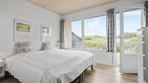 Ein Schlafzimmer mit einem großen Bett, einem Nachttisch, einem Fenster mit Vorhängen und zwei gerahmten Bildern an der Wand.