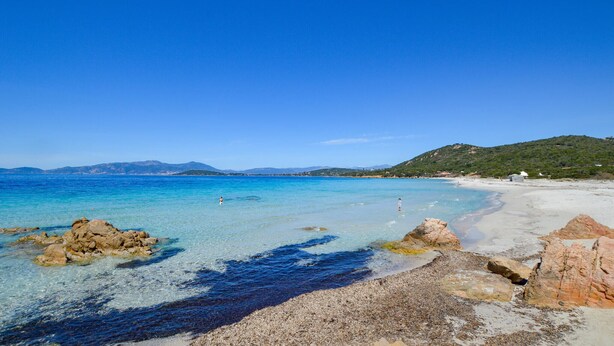 Una spiaggia con acque cristalline, formazioni rocciose e colline in lontananza.