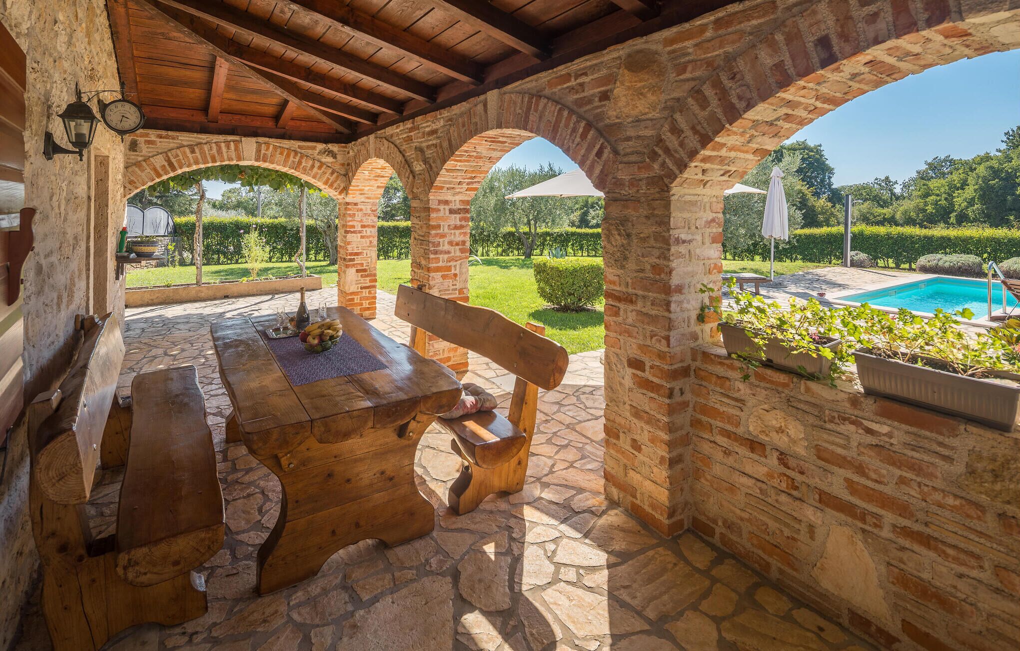 Un patio esterno rustico con un tavolo in pietra e panche, archi in mattoni e vista su una piscina.