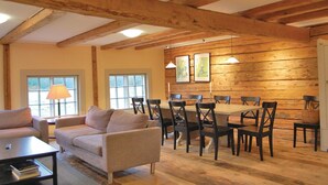 Un salon chaleureux avec un canapé, une table basse et un coin repas comprenant une table et des chaises. De grandes fenêtres offrent une vue sur l’extérieur, le plafond est orné de poutres en bois et les murs sont décorés de tableaux encadrés.