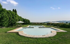 A rectangular swimming pool with a circular deck in the center, surrounded by grass and trees. There are lounge chairs and umbrellas in the background. - 3 bedroom beautiful home in Ischia di Castro -VT- (Ischia di Castro -VT-)