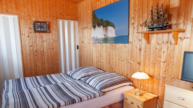 A wooden-paneled room with a bed, a nightstand, a desk, and a painting of a coastal scene. - Bungalow - Holiday Rentals Liliane (Göhren)