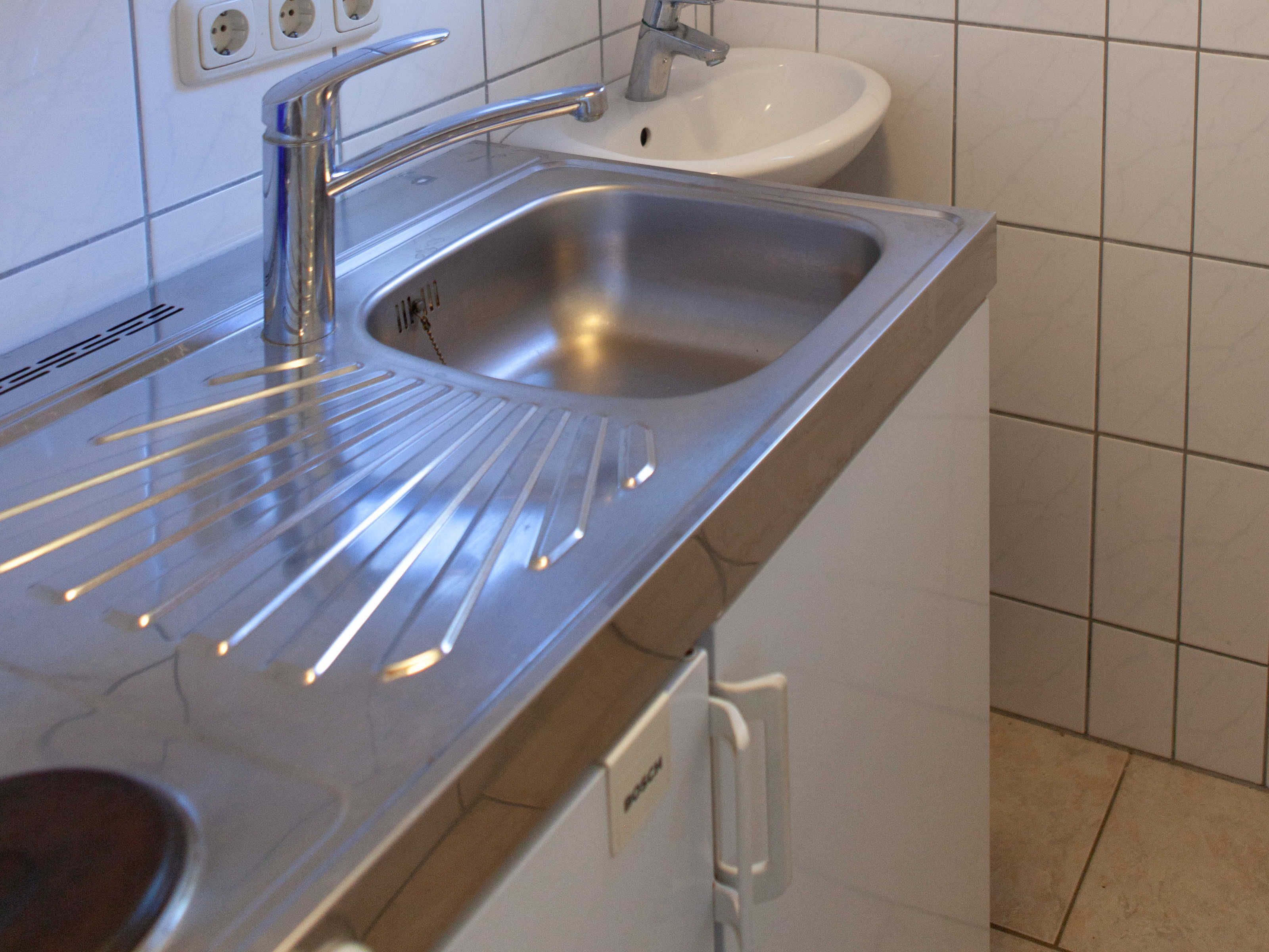 A stainless steel kitchen sink with a faucet, a white countertop, and a white wall with a tiled backsplash.