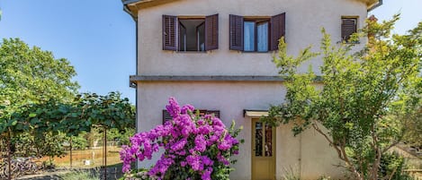 A two-story house with a yellow door, brown shutters, and a garden with purple flowers.