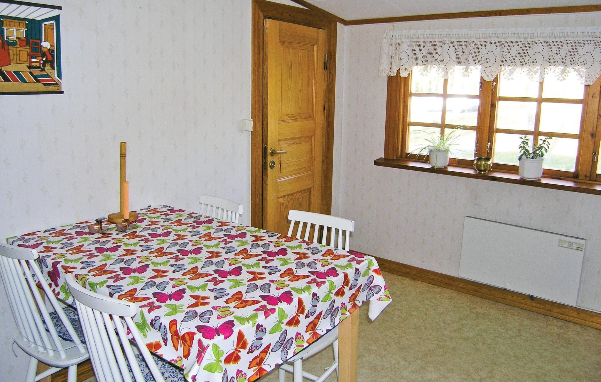 Comedor con una mesa cubierta por un mantel estampado con motivos de mariposas, sillas blancas, una puerta de madera y una ventana con plantas en macetas.