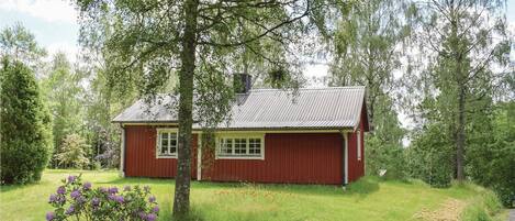 Ein rotes Holzhaus mit geneigtem Dach, umgeben von üppigem Grün und Bäumen.