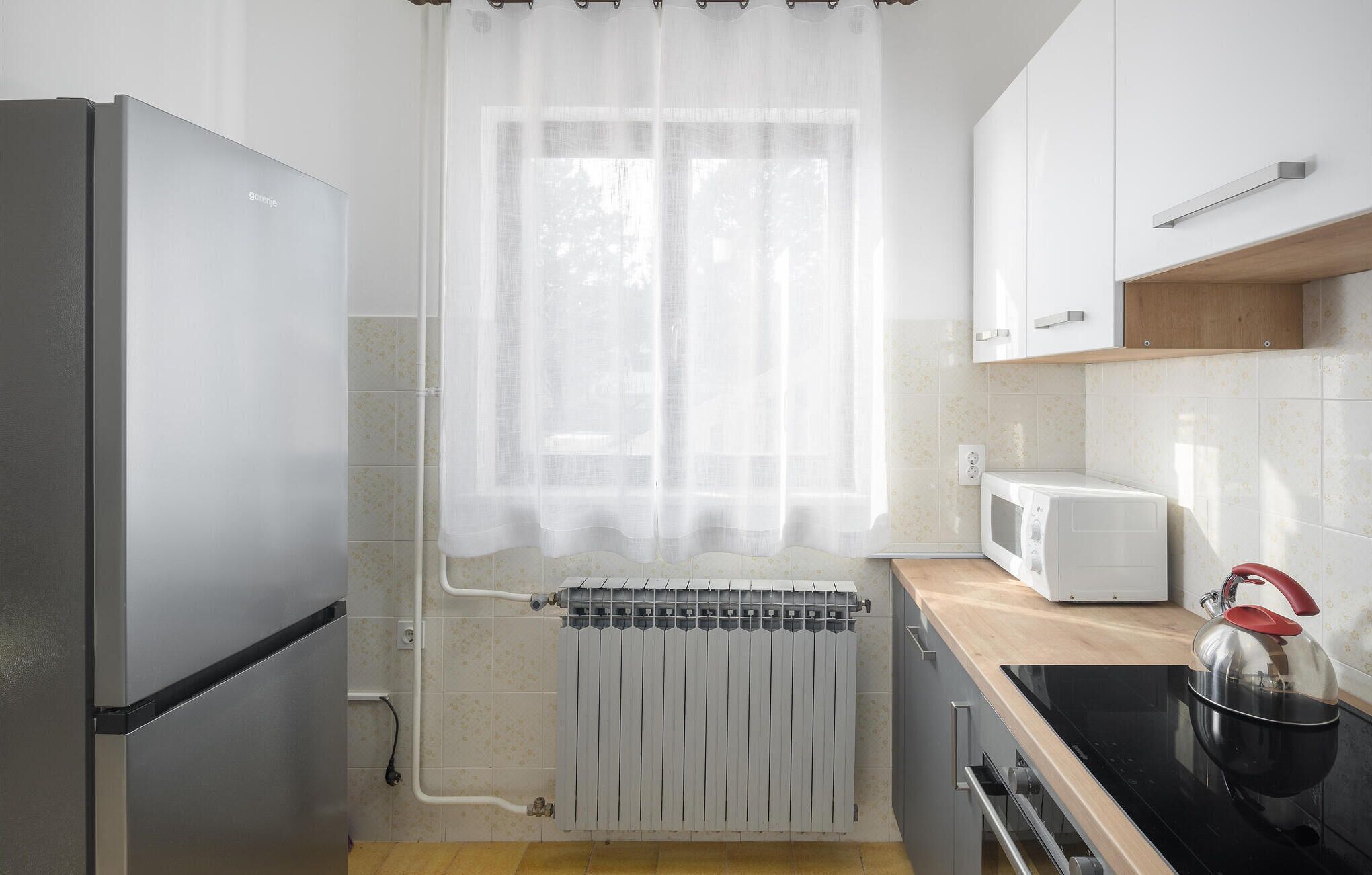 Eine kleine Küche mit Kühlschrank, Mikrowelle, Herd und einem Fenster mit transparenten Vorhängen.