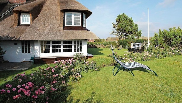 A thatched-roof house with a white exterior, a lawn with pink flowers, and a striped lounge chair.