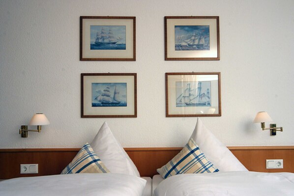 Habitación de hotel con dos camas, cada una con cabecera y almohadas. Cuatro cuadros enmarcados con motivos náuticos adornan la pared.