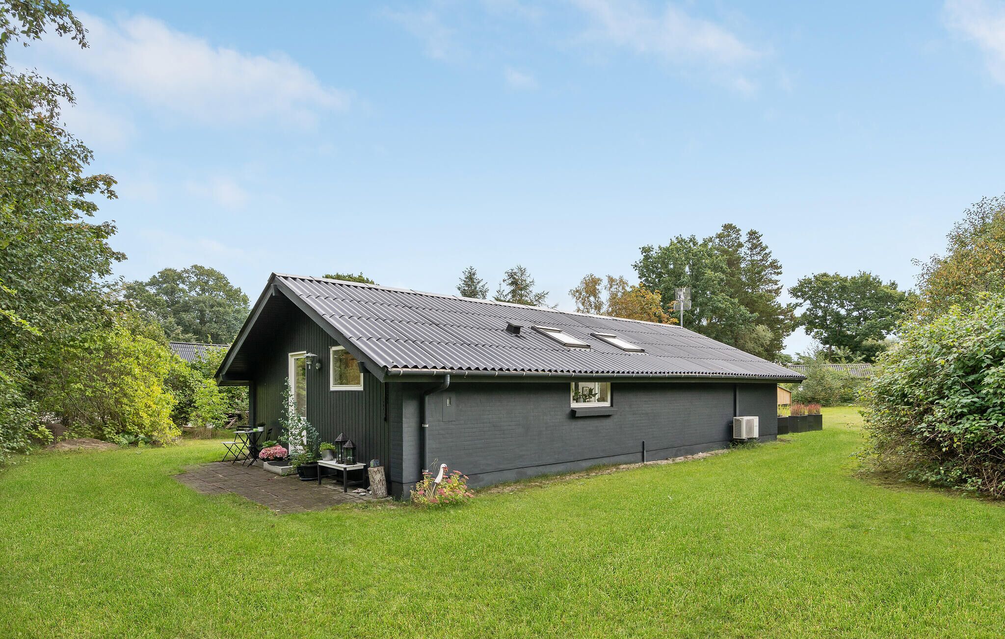 A small, dark-colored wooden house with a sloped metal roof, surrounded by a well-maintained lawn and various plants.