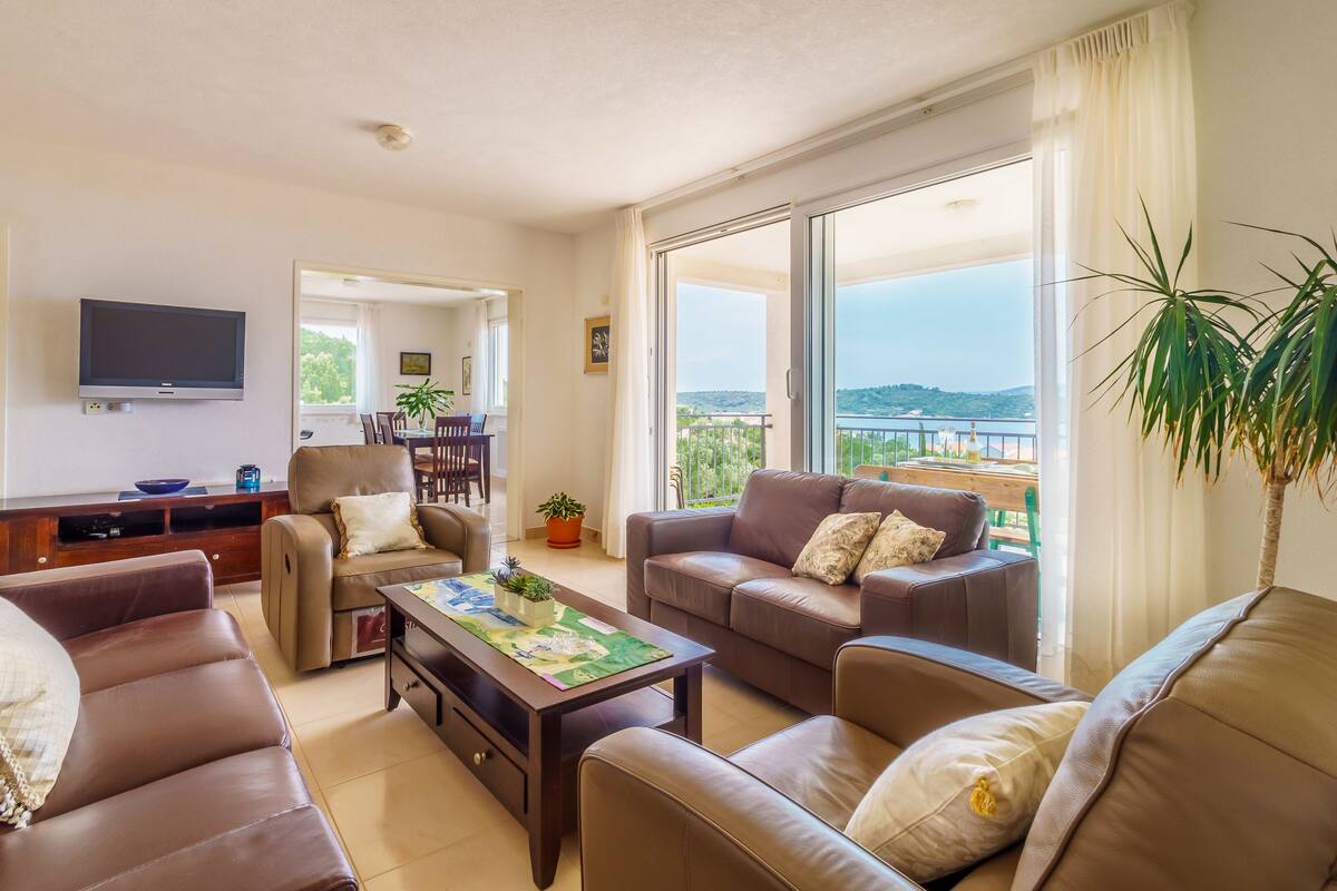 A spacious living room with leather sofas, a coffee table, and a TV. There is a balcony with a view of the sea.