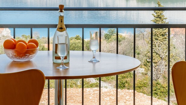 A table with a bottle and glasses, a bowl of oranges, and a view of a lake and mountains. - Kis Residence (Korcula)