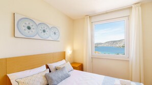 A neatly made bed with pillows, a bedside table with a lamp, and a window with a view of the sea. - Kis Residence (Korcula)