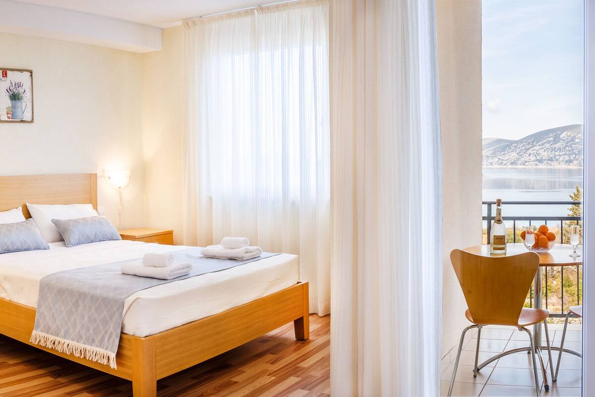 A bedroom with a wooden bed, a nightstand, a bedside table, and a view of a balcony with a table and chairs.