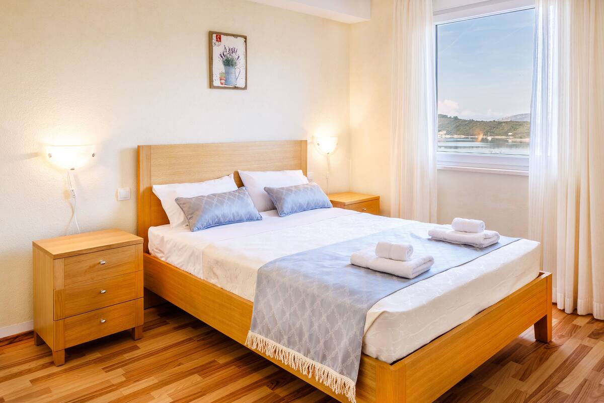 A neatly made bed with white linens and a blue blanket, wooden nightstands with drawers, a wall-mounted lamp, and a window with a view of a body of water and hills.