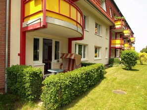 Un immeuble résidentiel à plusieurs étages doté de balcons, d’un petit patio avec une table et des chaises, et de haies bien entretenues.