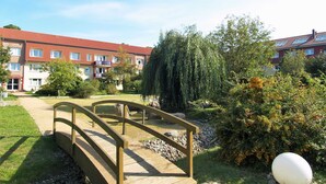 Eine Holzbrücke über einen Schotterweg, der zu einem Gebäude mit roten Ziegeldächern und vielen Fenstern führt.