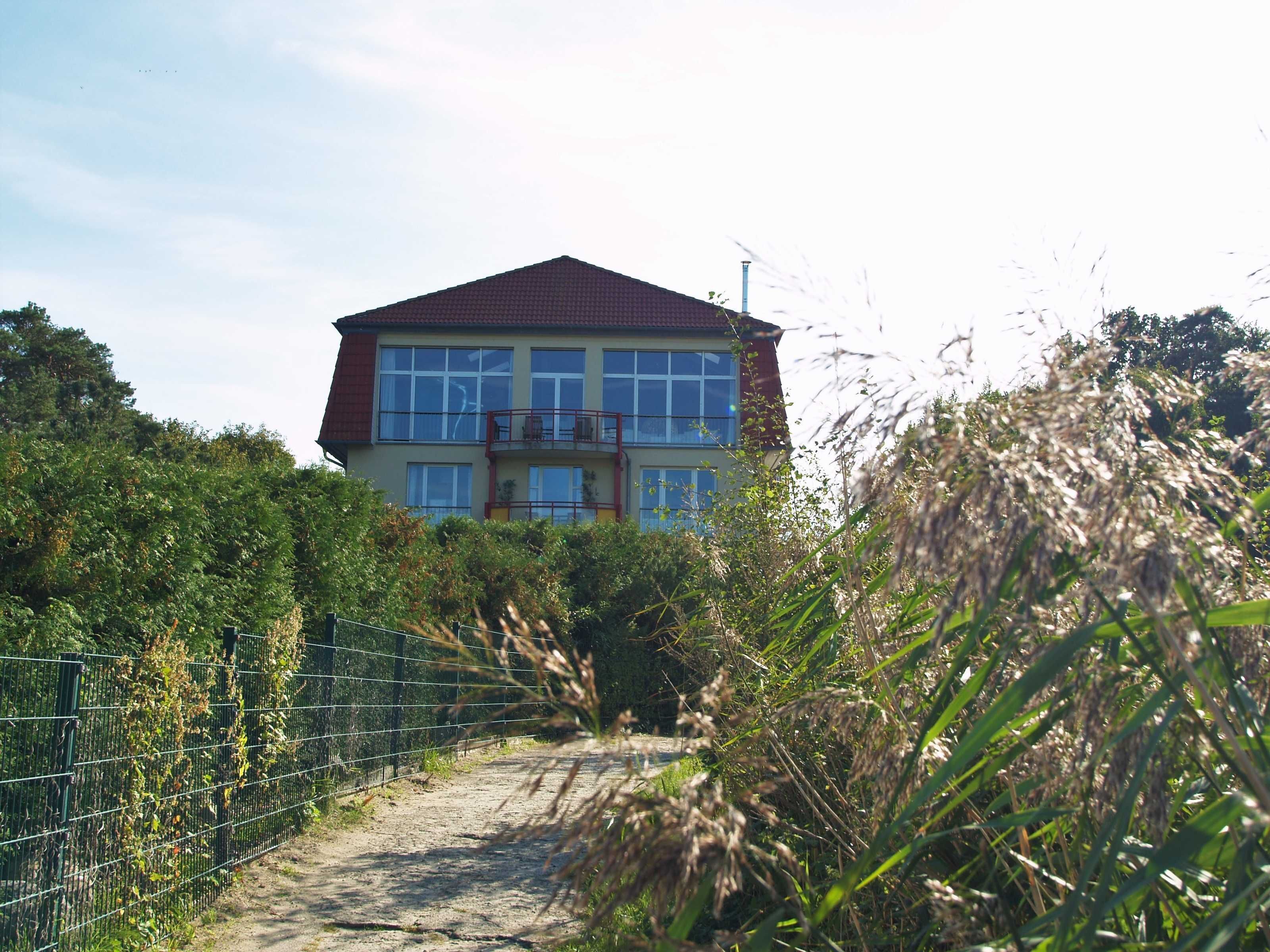A two-story house with a red roof, surrounded by dense vegetation and a metal fence.