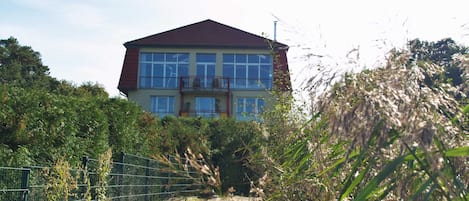 A two-story house with a red roof, surrounded by dense vegetation and a metal fence.