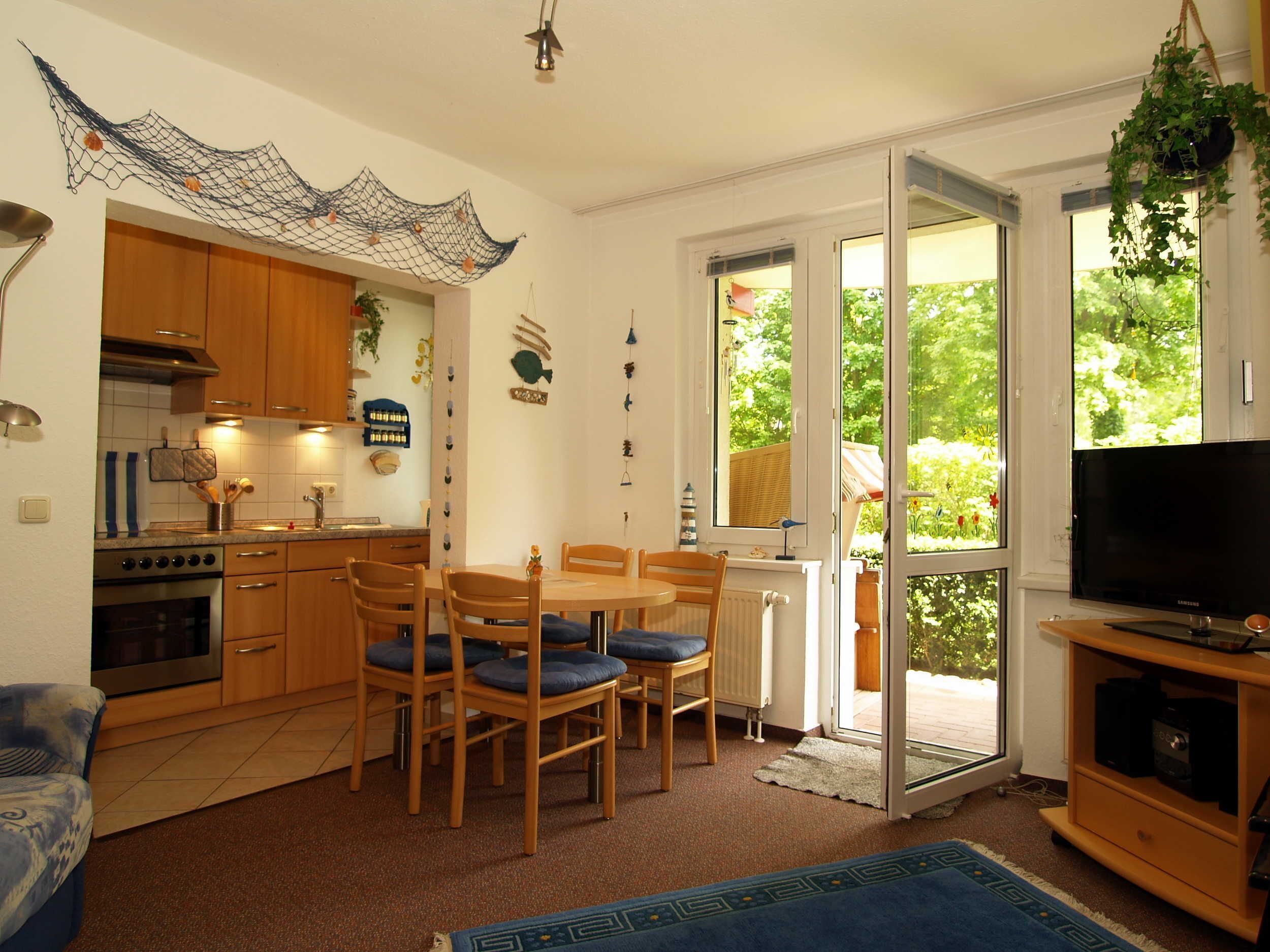 A compact kitchen with wooden cabinets, a dining table with chairs, a TV, and a large window with a view of greenery.