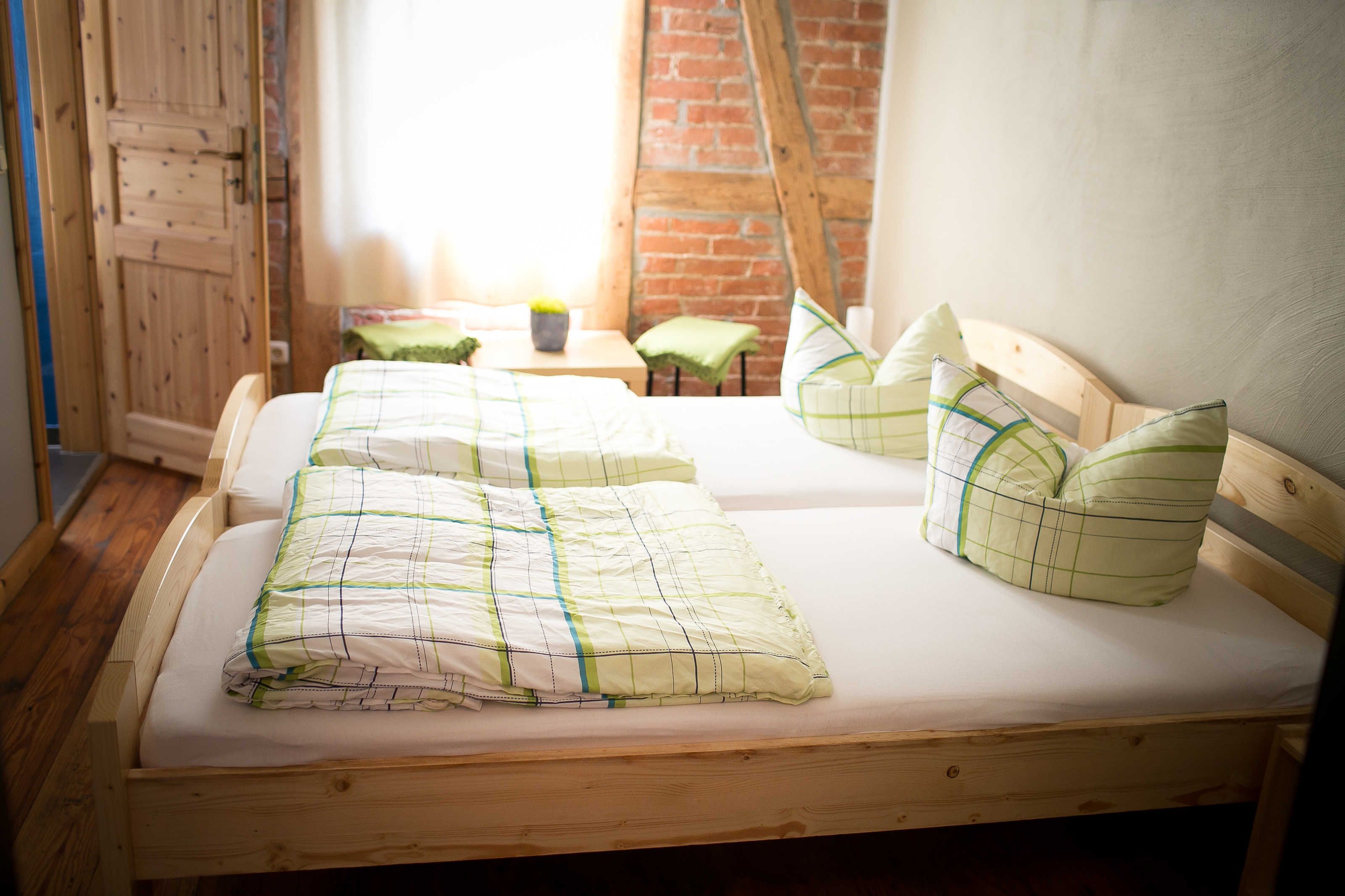 Ein hölzernes Bett mit einer grün-weißen Karossprenge und Kissen, ein kleiner Nachttisch mit einer Lampe und eine Backsteinwand im Hintergrund.