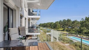 Un balcón moderno con barandilla de vidrio, mobiliario de mimbre y vista a una piscina y zona boscosa.