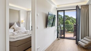 A hotel room with a bed, a TV, a balcony with a view of greenery, and a seating area. - Amazing apartment in Binz with outdoor swimming pool (Prora/Rügen)