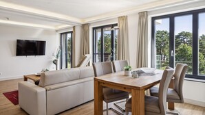 Una sala de estar moderna con un sofá, una mesa de comedor y sillas. Hay un televisor montado en la pared y ventanas grandes con cortinas.