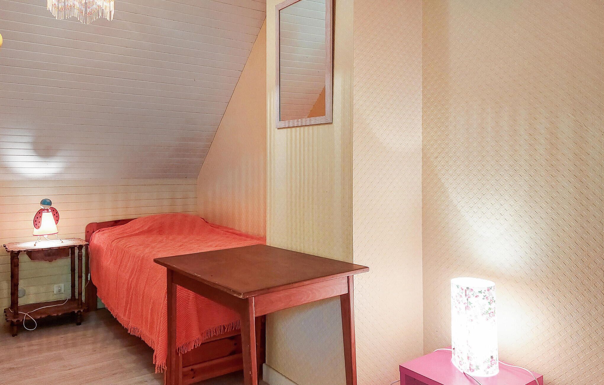 Un dormitorio con techo inclinado, una cama con manta roja, una mesita de noche de madera con lámpara y una mesita de noche rosa con lámpara floral.